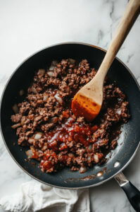 This image shows ketchup, brown sugar, vinegar, Worcestershire sauce, mustard, and salt being mixed into the browned beef and onion mixture, creating a tangy and flavorful filling.