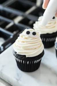 A black buttercream base is being piped onto a cooled cupcake on the white marble cooktop. Thin white buttercream lines are added for mummy bandages, with candy eyes placed on top