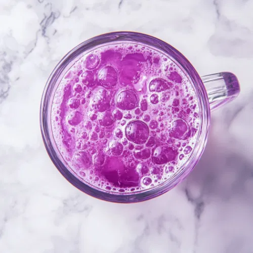 The completed grape lemonade drink, served in the glass cup over the white marble cooktop. The fizzy purple drink is ready to be enjoyed, with bubbles gently rising to the surface.