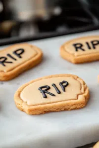 A close-up of Milano cookies being halved on the white marble cooktop, with black gel icing used to write 'RIP' on each tombstone-shaped cookie for decoration.