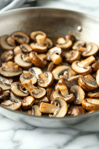 This image shows mushrooms being sautéed in a pan, turning golden brown with a slight sizzle, ready to be layered on top of the polenta.