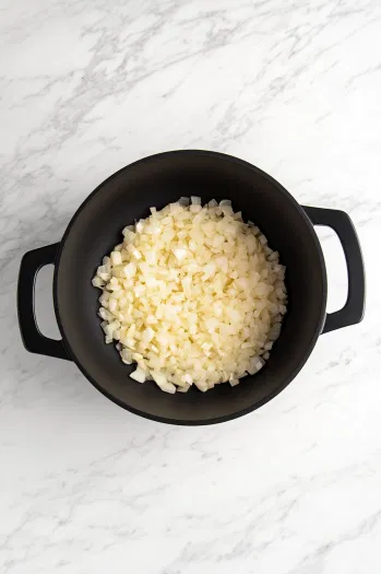 The black pot on the white marble cooktop contains olive oil heated with diced onions sautéing for about 5 minutes. The onions are translucent, and minced garlic is being added for an additional 1-2 minutes of cooking.