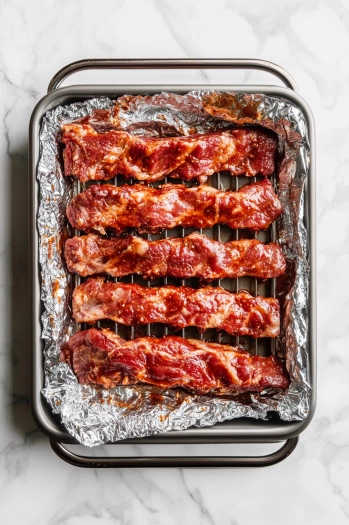 The ribs are placed in a roasting pan with a rack on the white marble cooktop. They are slathered with keto BBQ sauce, and aluminum foil is tented over the top to keep the moisture in during roasting.