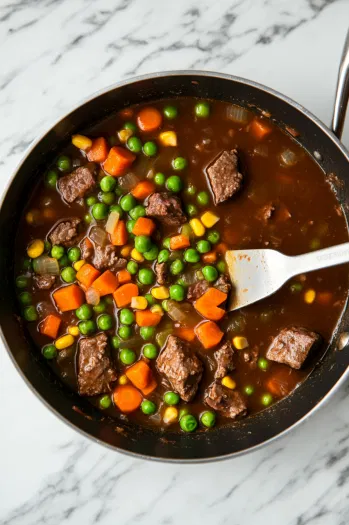 This image shows beef broth being added to the meat mixture, followed by frozen peas, carrots, and corn simmering together in the skillet.