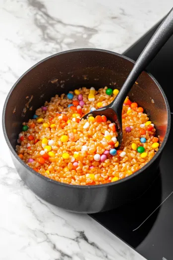 Rice Krispies cereal is stirred into the melted marshmallow mixture in the black saucepan over the white marble cooktop. Halloween M&Ms, sprinkles, and candy corn are gently folded in for even distribution