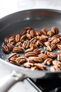 This image shows pecans being toasted in a skillet over medium-low heat, with constant stirring to prevent burning.