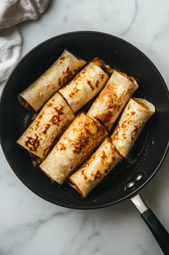 This image shows the rolled tortillas placed seam-side down in a skillet, toasting to a golden brown and crispy exterior.