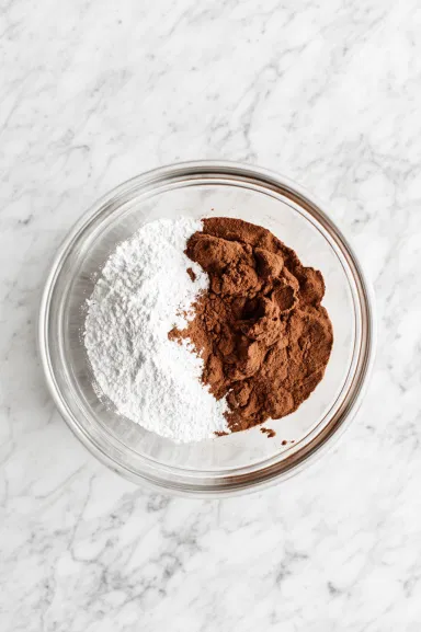 Baking powder, salt, and pumpkin pie spice are added to the well-blended wet ingredients in a glass bowl sitting on the white marble cooktop, ready to mix with the dry ingredients.