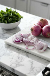 This image shows onions being peeled and thinly sliced on a cutting board. A sharp knife is being used to create even slices of onion.