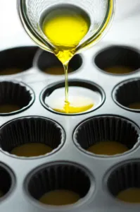 A teaspoon of vegetable oil is poured into each cup of a 12-cup muffin tin resting on the white marble cooktop, with oil evenly distributed across all the cups.