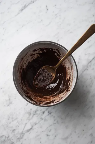 The mixing bowl on the white marble cooktop as cocoa powder and red wine vinegar are whisked in. Grated dark chocolate is gently folded in with a metal spoon, resulting in a rich, chocolatey meringue mixture.