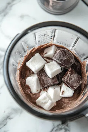 The blender on the white marble countertop with frozen sweetened cream cubes and melted chocolate being added, blending into a rich, smooth mixture.