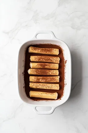 The baking dish on the white marble cooktop, with a smooth layer of mascarpone mixture spread over the soaked ladyfingers, topped with an even dusting of cocoa powder for a rich chocolate layer.