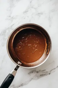 The saucepan remains on the white marble cooktop with the heat turned off. Oats, peanut butter, and vanilla extract are stirred in, creating a thick and creamy mixture with a satisfying texture, ready for forming into cookies.