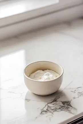 A small cup on the white marble cooktop containing a mixture of white wine vinegar and cornflour. This stabilizing mixture is gently folded into the glossy meringue.