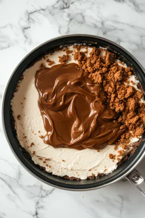 A small pan on the white marble cooktop holds Biscoff spread melting gently on low heat until it reaches a pourable consistency. The chilled cheesecake in its tin awaits as the melted spread is poured over the top, with crushed Biscoff biscuits sprinkled around the edges for a decorative finish.