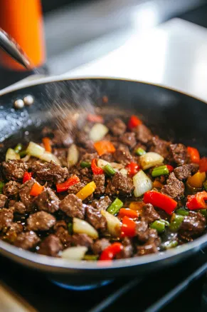 Minced garlic is being added to the browned beef and vegetables in the skillet on the white marble cooktop, creating a savory aroma.