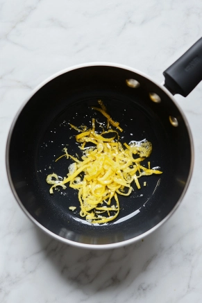 A black saucepan on a white marble cooktop, with finely diced lemon peel being added to the gently simmering water and sugar mixture, releasing aromatic steam.