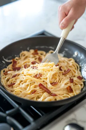 Cooked pasta is being folded into the creamy sauce in the skillet over the white marble cooktop, followed by the addition of Monterey Jack cheese and crispy bacon pieces.