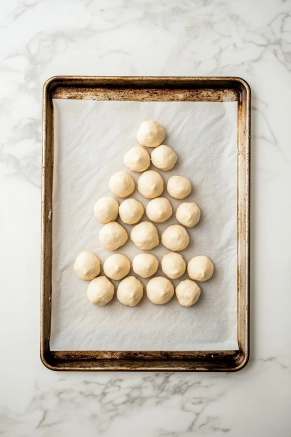 A parchment-lined baking sheet on the white marble cooktop holds the dough balls arranged seam-side down in a Christmas tree shape. The balls touch one another, creating a festive design.