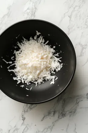 The gelatin mixture is transferred to a black mixing bowl on the white marble cooktop and beaten until smooth. Vanilla extract and flaked coconut are ready to be folded in.