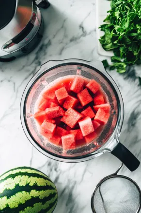 A blender on the white marble countertop filled with watermelon cubes being blended into smooth juice, with a strainer nearby for removing seeds and pulp.