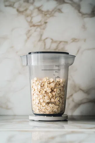 A sleek blender on a white marble cooktop filled with rolled oats, water, maple syrup, vanilla extract, and a pinch of sea salt, with the lid closed and the mixture blending into a creamy oat milk.