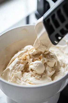 The chilled mixture is poured into an ice cream maker on the white marble cooktop, churning to a soft-serve consistency. Finely chopped crystallized ginger is added for extra flavor and churned briefly to combine.