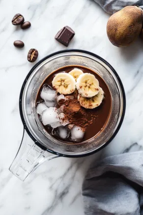 A blender on a white marble countertop filled with almond milk, cold coffee, dark cocoa powder, a frozen banana, and ice, ready to blend.