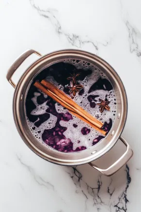 A large pot on the white marble countertop filled with purple corn, water, a cinnamon stick, and whole cloves, beginning to boil.