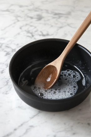 A microplane grater zesting a fresh lemon over a small black bowl on a white marble cooktop, with the finely diced yellow peel neatly collected in the bowl.