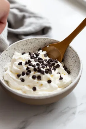 A bowl on a white marble counter with ricotta cheese, whipped cream, vanilla, and chocolate chips being gently folded together into a smooth mixture.