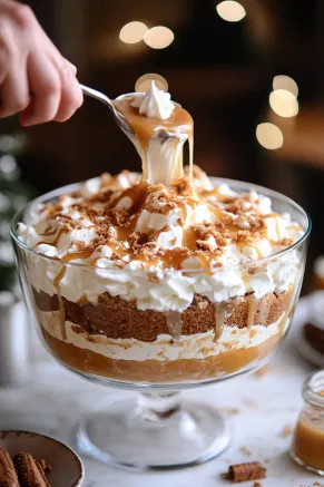 The trifle bowl on the white marble cooktop is gradually filled with more layers of gingerbread cake, alternating with plain custard, Biscoff custard, whipped cream, and caramel until the bowl is filled to the top. The final layer is finished with a smooth, even layer of whipped cream.