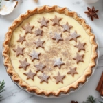 The finished tart rests on the white marble cooktop, its surface adorned with pastry stars lightly dusted with icing sugar and cinnamon. The tart’s creamy custard filling and festive decorations make it ready to be sliced and enjoyed.