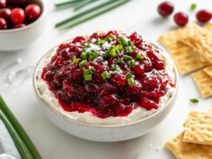 The strained cranberry mixture layered over the cream cheese base on a serving platter, garnished with green onion greens, flaky sea salt, and a drizzle of honey, accompanied by crackers on the white marble cooktop.