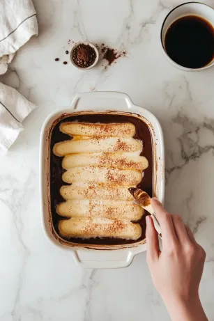 A hand dipping each ladyfinger into the espresso mixture on the white marble cooktop, holding each side in the liquid for a count of three before layering them neatly in the baking dish.