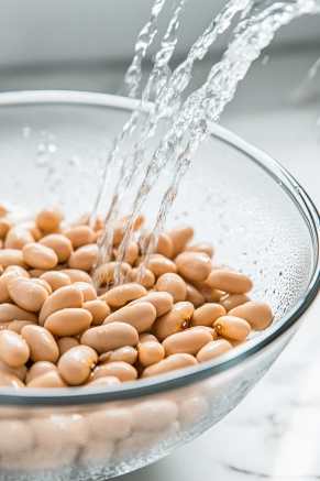 Soaked Great Northern beans are being drained from a glass bowl into a colander on the white marble cooktop, with water streaming from the beans as they are rinsed thoroughly.