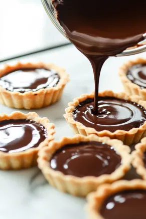 Chocolate ganache being poured into cooled mini tart shells on the white marble countertop, evenly filling each one.