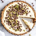 A chilled cannoli cream pie topped with mini chocolate chips and chopped pistachios, neatly sliced with a glossy finish, displayed on a clean white marble background.