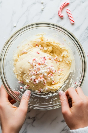 A glass mixing bowl on the white marble cooktop is filled with cookie dough, as white chocolate chips and crushed candy canes are being gently folded in, adding festive flair.