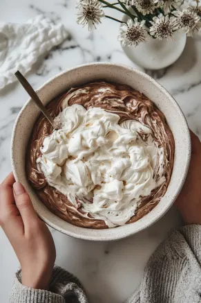 The cream cheese mixture in a large bowl on the white marble cooktop as a portion of the whipped cream is gently folded in, lightening the filling. The remaining whipped cream is folded in two stages until fully combined and fluffy.