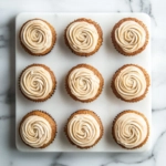 The cooled cupcakes are displayed on the white marble cooktop, each topped with a swirl of cream cheese frosting. The spiced cupcakes are ready to be stored in the refrigerator with a reminder to bring them to room temperature before serving for the best flavor.