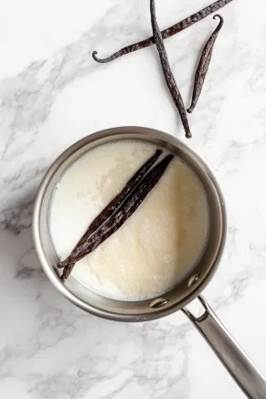 A saucepan on the white marble cooktop contains heavy cream, granulated sugar, and vanilla bean seeds with the pod. The mixture is heated gently, dissolving the sugar.