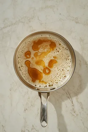 A saucepan on the white marble cooktop heats cream, milk, and ginger syrup, with small bubbles forming at the edges, indicating it has reached the scalding point.