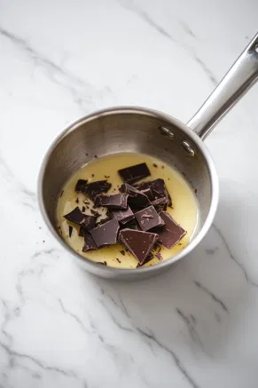 A small saucepan on the white marble cooktop with honey and sugar gently heating. The mixture simmers on low for 3 minutes until the sugar dissolves, then dark chocolate is added, melting smoothly into a rich, syrupy base.