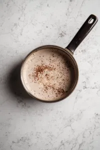 The saucepan remains on the white marble cooktop with the heat turned off. Oats, peanut butter, and vanilla extract are stirred in, creating a thick and creamy mixture with a satisfying texture, ready for forming into cookies.