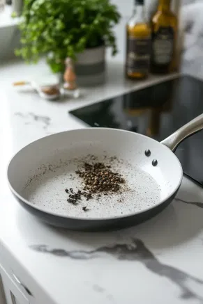 A small dry pan on a white marble cooktop with crushed spices being gently heated for 1-2 minutes. The heat enhances the deep, spicy aromas, preparing the spices for the gin infusion.