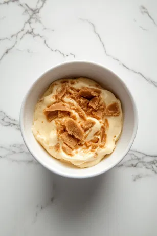 A microwave-safe bowl on the white marble cooktop holds half of the custard combined with Biscoff spread. The mixture is warmed in 30-second intervals, being stirred each time until the Biscoff melts smoothly into the custard, creating a rich layer for the trifle.