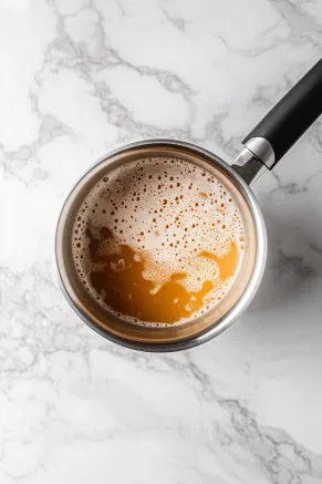 A saucepan on the white marble countertop with butter and brown sugar melting together, bubbling as condensed milk is whisked in to create a thick caramel.