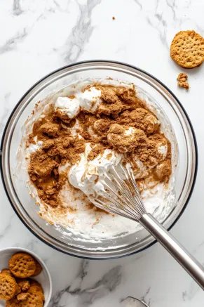 A large mixing bowl on the white marble cooktop holds double cream, cream cheese, and Biscoff spread. An electric whisk blends the ingredients into a thick, creamy mixture with an ice-cream-like consistency, ready to layer over the biscuit base.